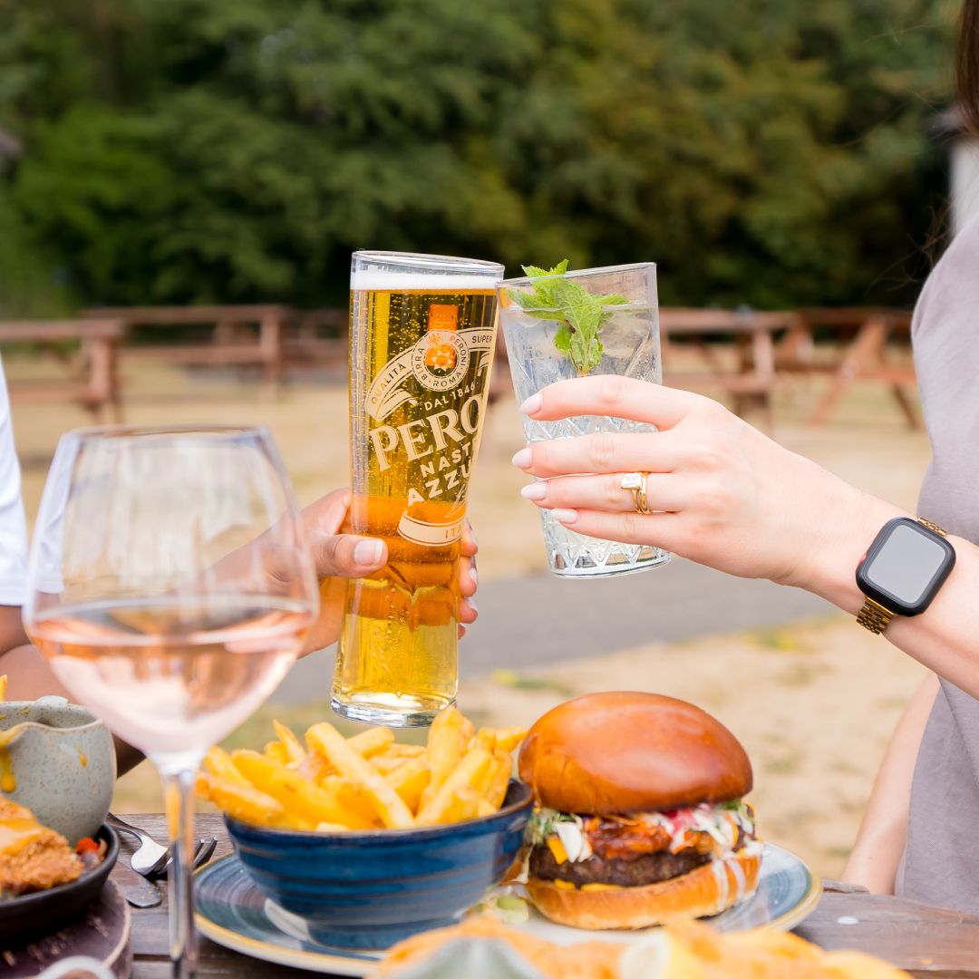 A zoomed-in photo of two people sitting at a wooden picnic bench in a large UK beer garden. There is a selection of food and drink on the table, including a loaded burger, and the people are holding full glasses and clinking them in a cheers motion. One person holds a glass of Peroni, and the other holds a soft drink.